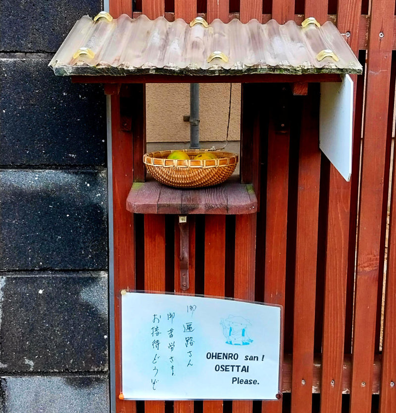 A small bowl of oranges set in a covered hatch in a fence along the Shikoku Henro trail